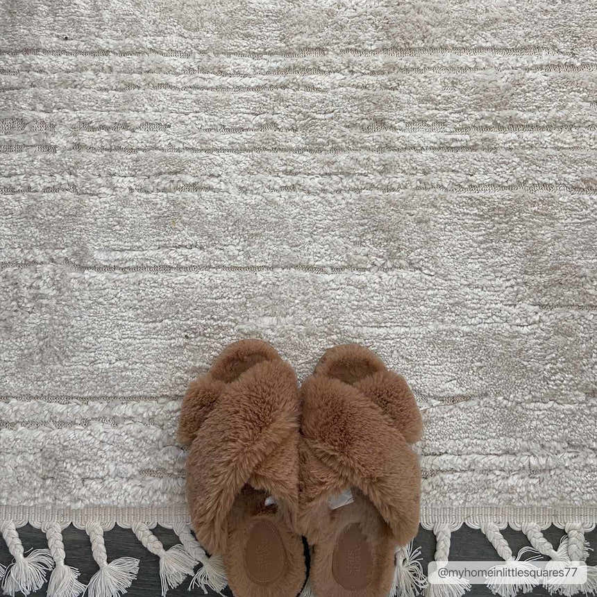 Close-up of the textured ivory Ibarra rug with tassels, paired with fuzzy brown slippers on a dark wood floor. Styleshot