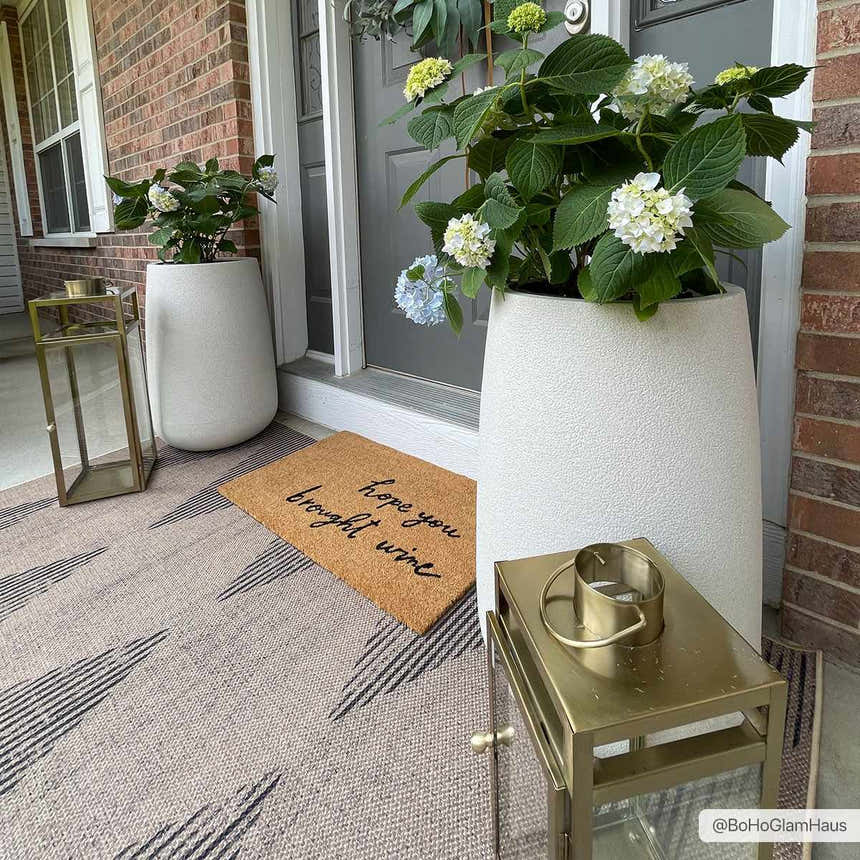 Front porch scene with the taupe Ijlal Washable Area Rug, potted hydrangeas, gold lanterns, and a funny doormat. Roomscene-Rectangle