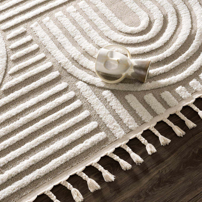Close up of the Moise Area Rug featuring a beige background and cream geometric pattern with tassels. Styleshot