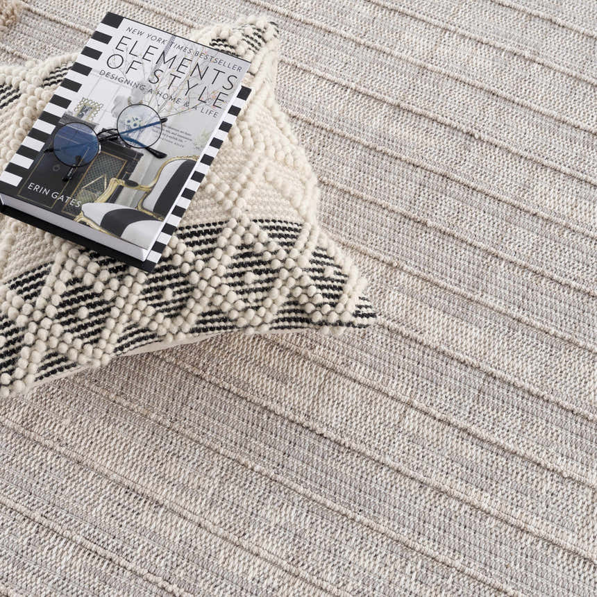 Close-up of the Sulak Cream Area Rug showing its textured, striped design, paired with a book and patterned pillow. Styleshot