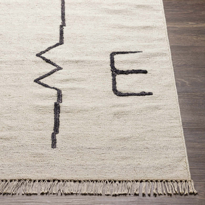 Close-up of the hand-woven AISHA Area Rug. Beige wool rug with a black geometric pattern and fringe. Contemporary style. Front
