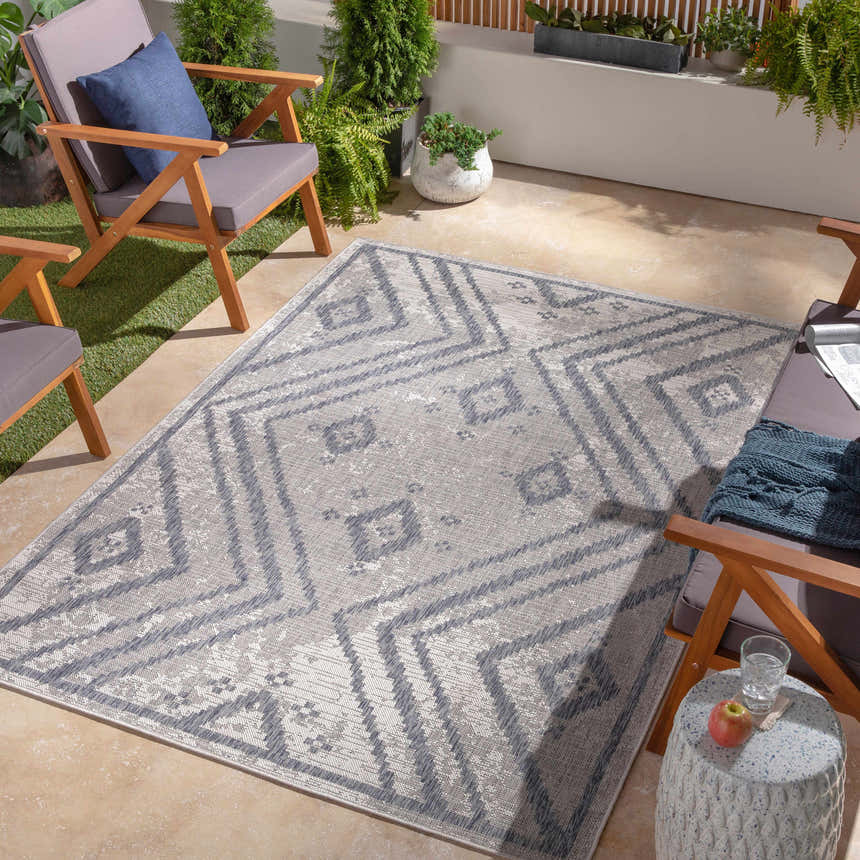 Outdoor patio scene with Auckland Indoor & Outdoor Rug in dark gray and natural gray, surrounded by wood chairs and lush greenery. Roomscene-Rectangle