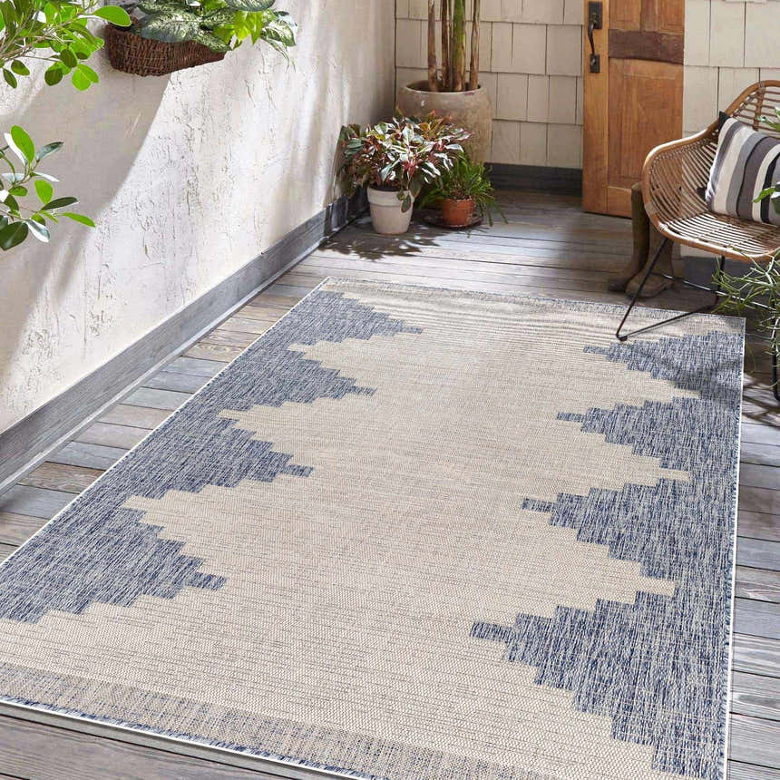 Bohemian patio scene with a Djugun Blue Outdoor Rug, wicker chair, and potted plants. The rug features a geometric blue and oatmeal pattern. Roomscene-Rectangle