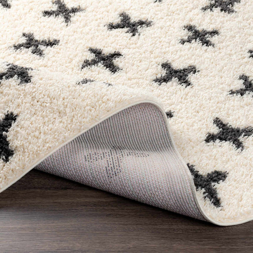 Close-up of the Gambo Area Rug. Cream shag rug with a bold black plus sign pattern. Machine woven polypropylene. Fold