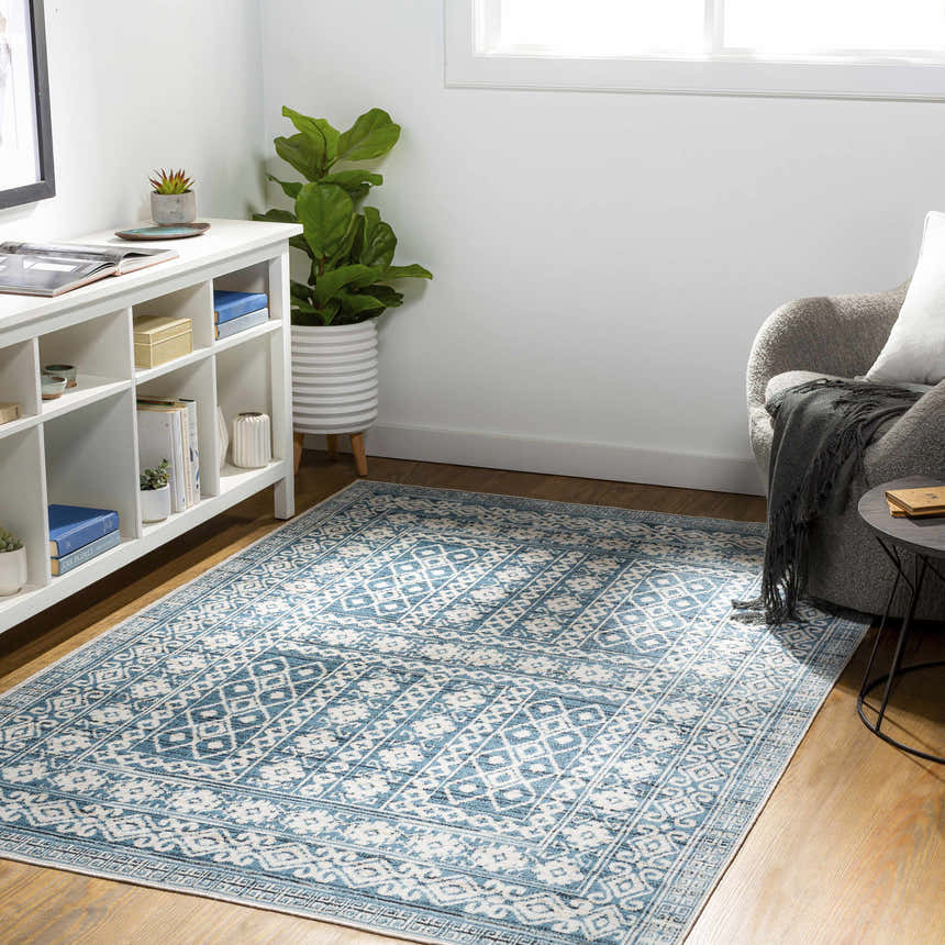 Bright living room scene with blue and cream Gandy Washable Area Rug, white shelving, potted plant, and gray armchair. Roomscene-Rectangle