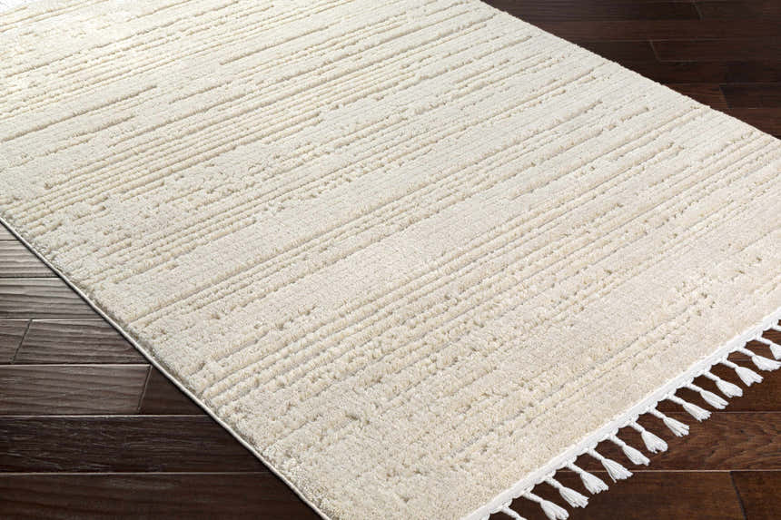Close-up of the ivory Ibarra Textured High/Low Pile Rug with Tassels, showing texture and tassel detail on dark wood floor. Corner