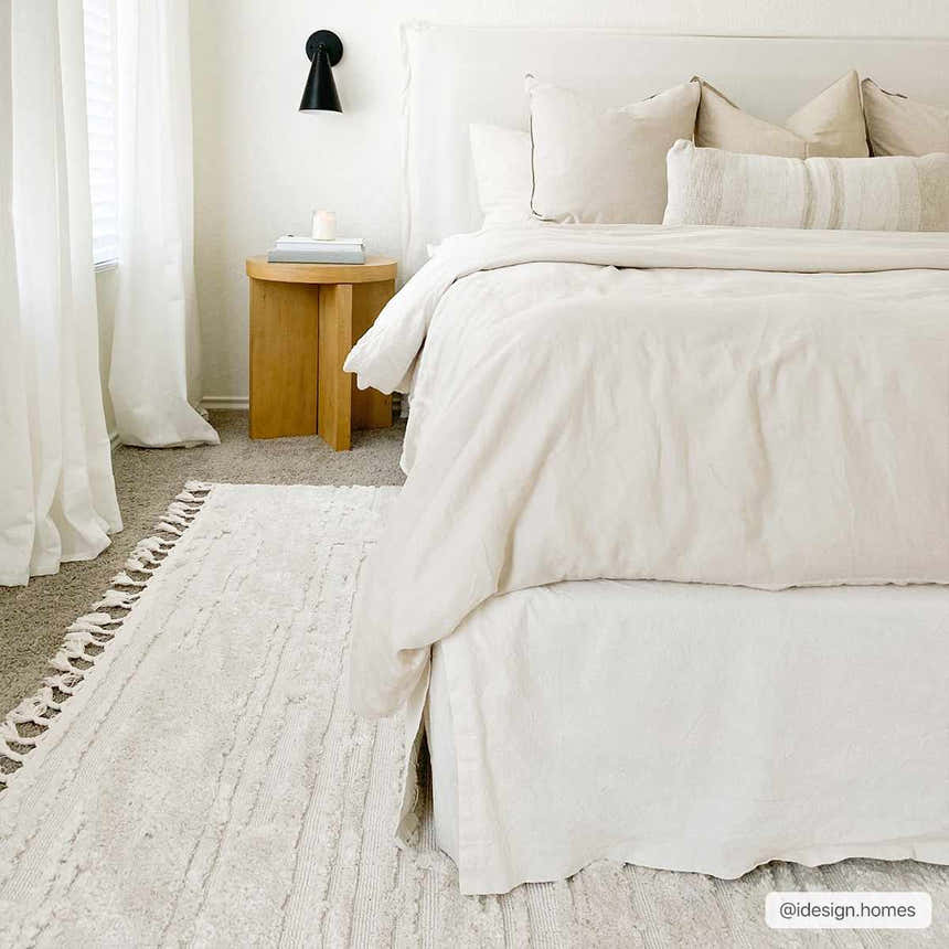Bedroom scene with the Ibarra Textured Ivory Rug, white bedding, and a wooden side table. Neutral, cozy aesthetic. Roomscene-Rectangle