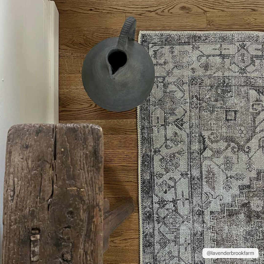 Close-up of Athor Washable Rug in dark gray, eggplant, and natural gray on wood floor with rustic wood stool and gray ceramic vase. Styleshot