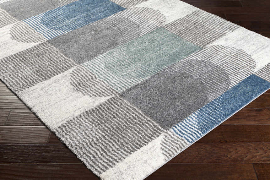 Close-up of the Melber Area Rug showing a geometric block pattern in shades of blue, gray, and white on a dark wood floor. Corner