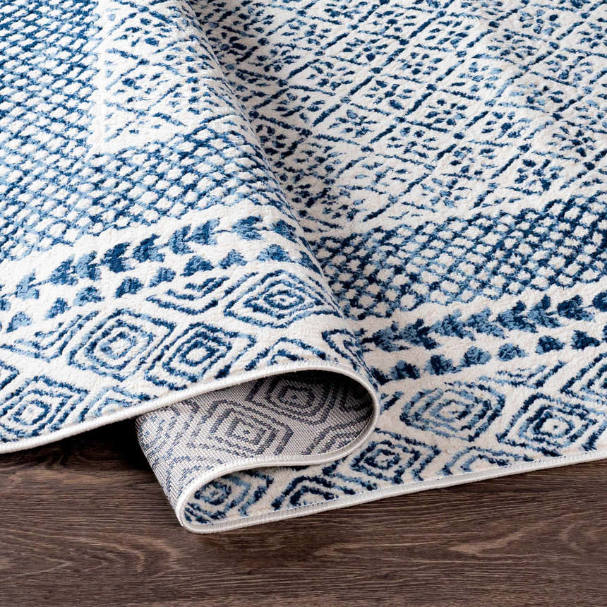 Close-up of the Mohrsville Area Rug featuring a blue and white geometric pattern, rolled on a dark wood floor. Fold
