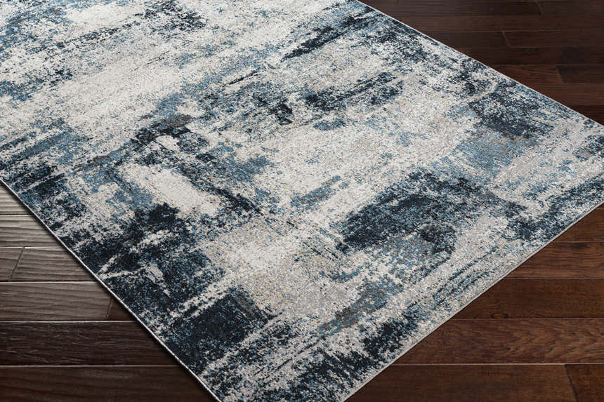 Close-up of Pugo Washable Area Rug in blue, cream, and gray on dark wood floor. Abstract contemporary pattern. Corner