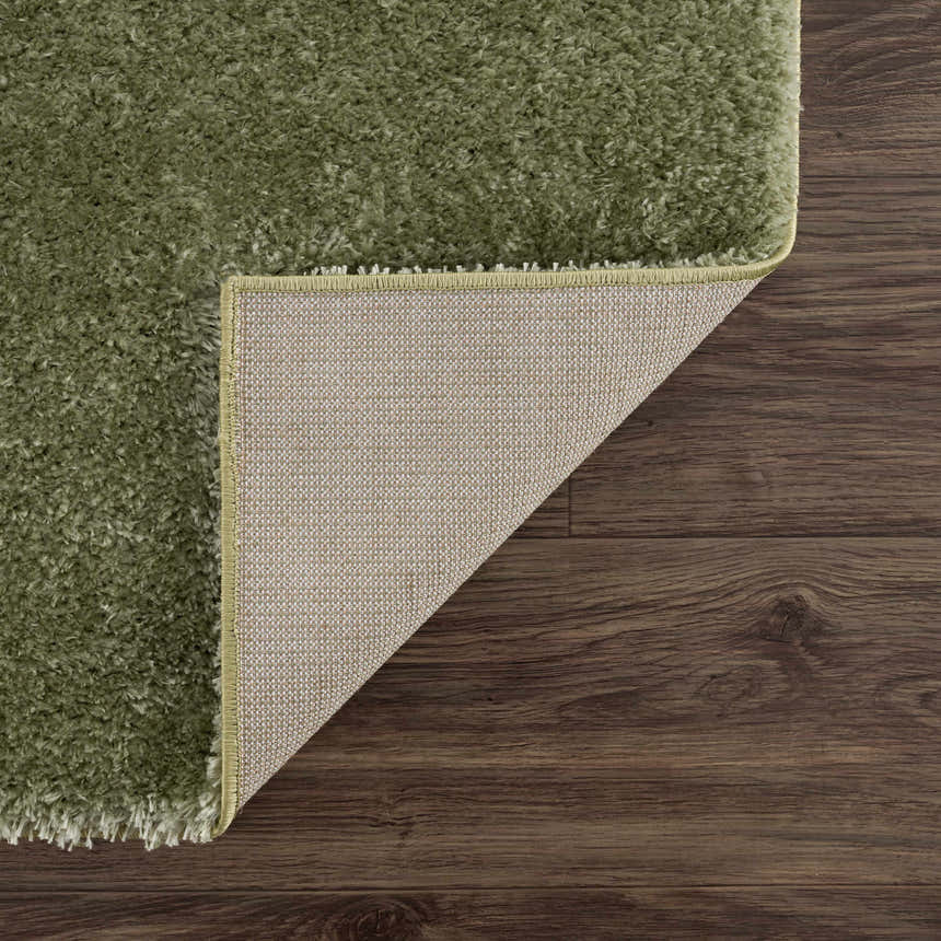Close-up of the Heavenly Solid Green Plush Rug showing the shag texture and rug backing on dark wood floor. Fold