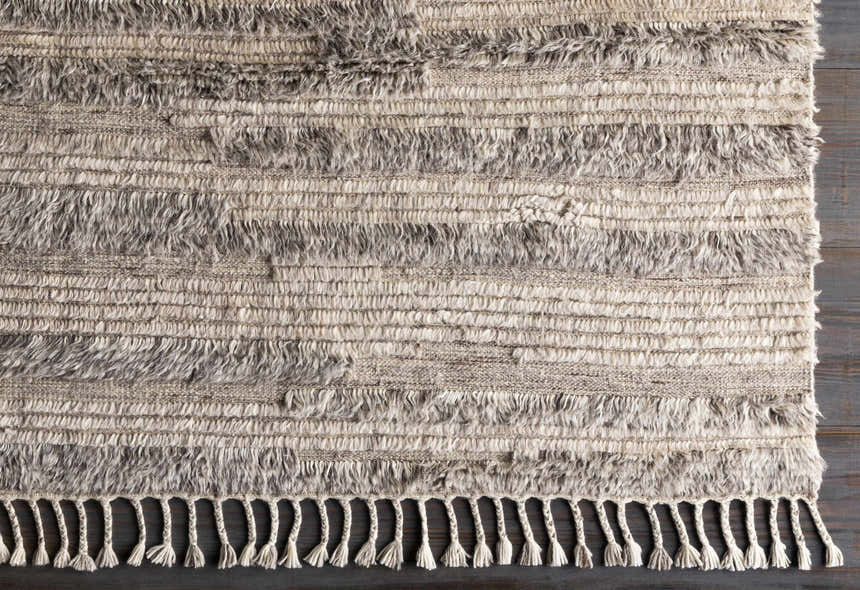 Close-up of the hand-knotted Saundersfoot Area Rug. Charcoal, gray, and light beige wool with a textured, bohemian design and fringe. Corner