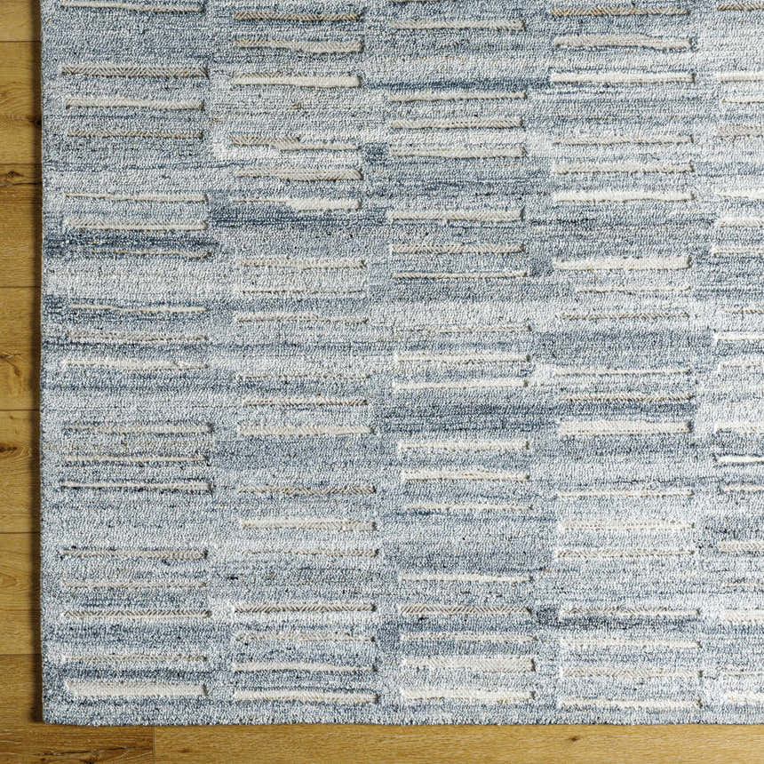 Close-up of the hand-tufted Darcy Area Rug. Features a textured, geometric pattern in shades of blue, white, and tan. Corner