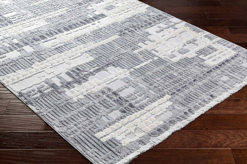 Close-up of Bodhi Textured Saxony Rug with Fringes. Gray, light gray, and white geometric pattern. Fringed edge detail. Corner