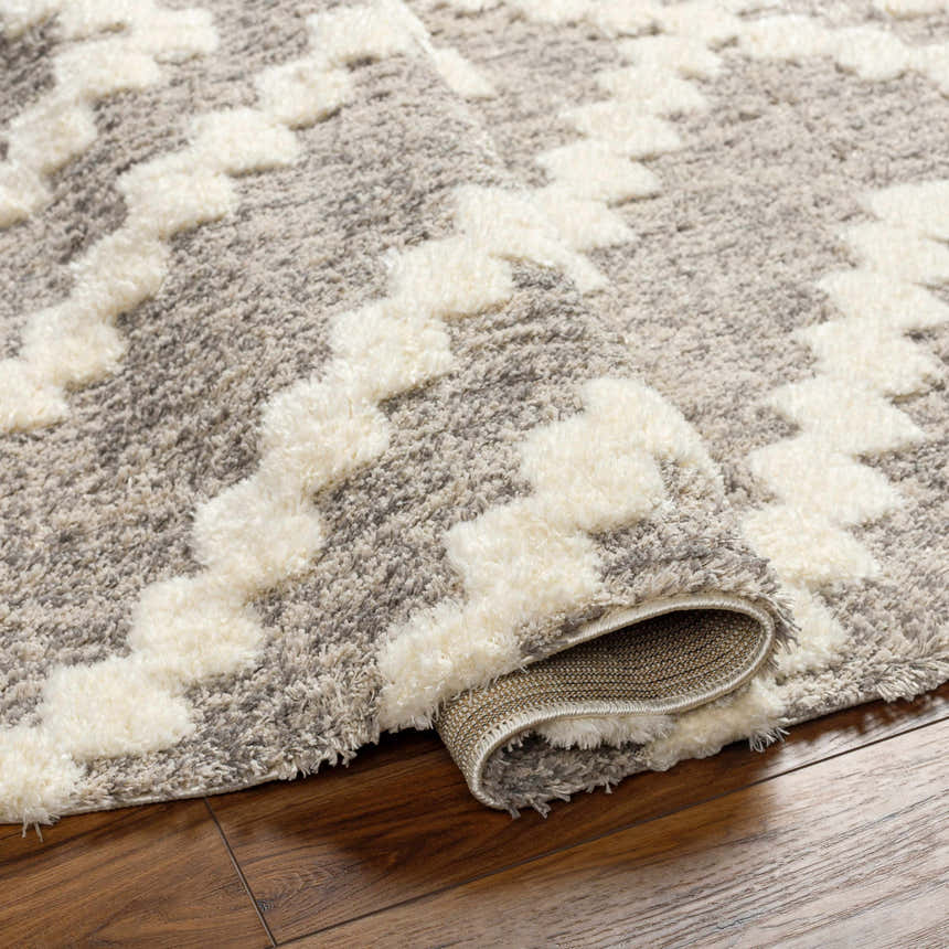 Close-up of the Rian Beige Area Rug showing its shag texture and bone geometric pattern on a gray background. Fold