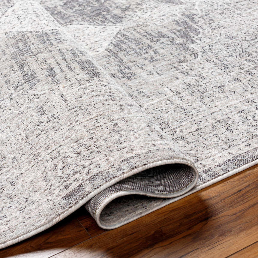 Close-up of the Azibo Area Rug showing its light gray, taupe, and black traditional pattern and recycled fabric texture. Fold