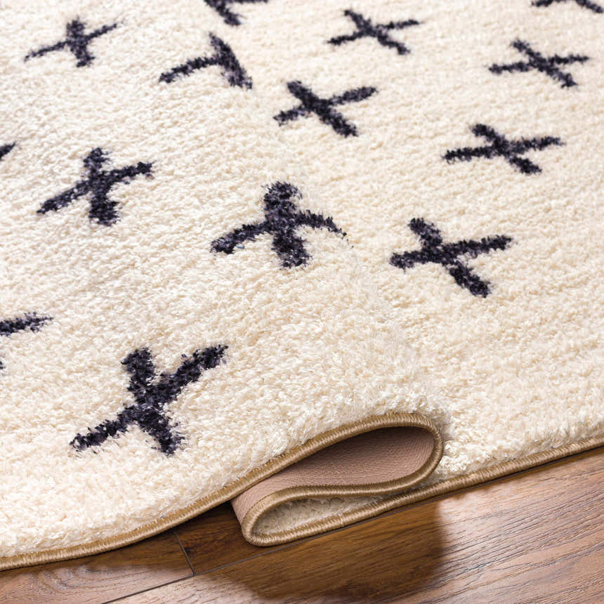 Close-up of the Bede Washable Area Rug. Beige shag rug with charcoal X pattern and bound edge. Polyester material. Fold