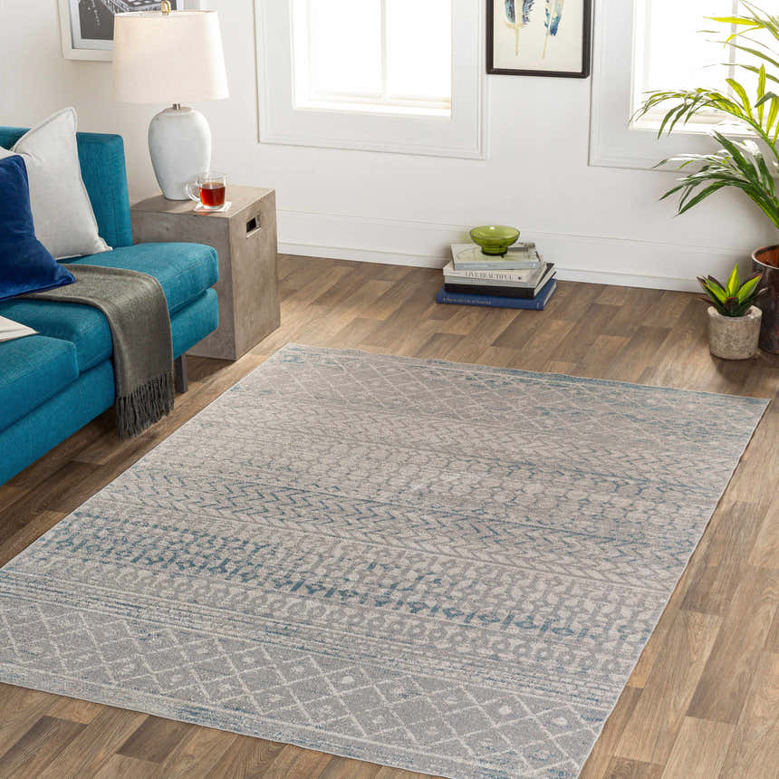 Living room scene with Tambac Blue & Gray Washable Area Rug, teal sofa, wood floors, and bright natural light. Roomscene-Rectangle