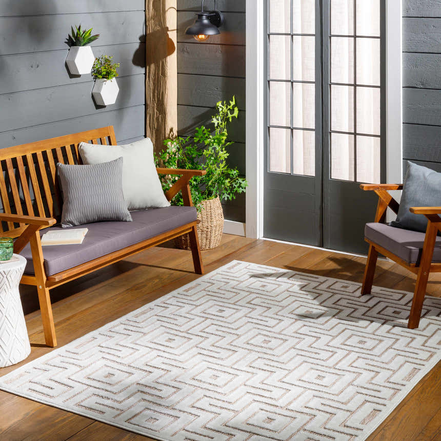 Cozy patio scene with a Zevi Tan Outdoor Rug, wood furniture, grey cushions, and plants against a grey wood wall. Roomscene-Rectangle
