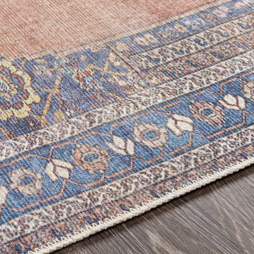 Close-up of the Stakeford Washable Area Rug showing light blue, navy, orange, and tan colors with a detailed traditional pattern. Texture