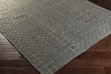 Close-up of the gray Sunburst Area Rug. Hand-woven leather rug with a basketweave pattern on dark wood floors. Corner
