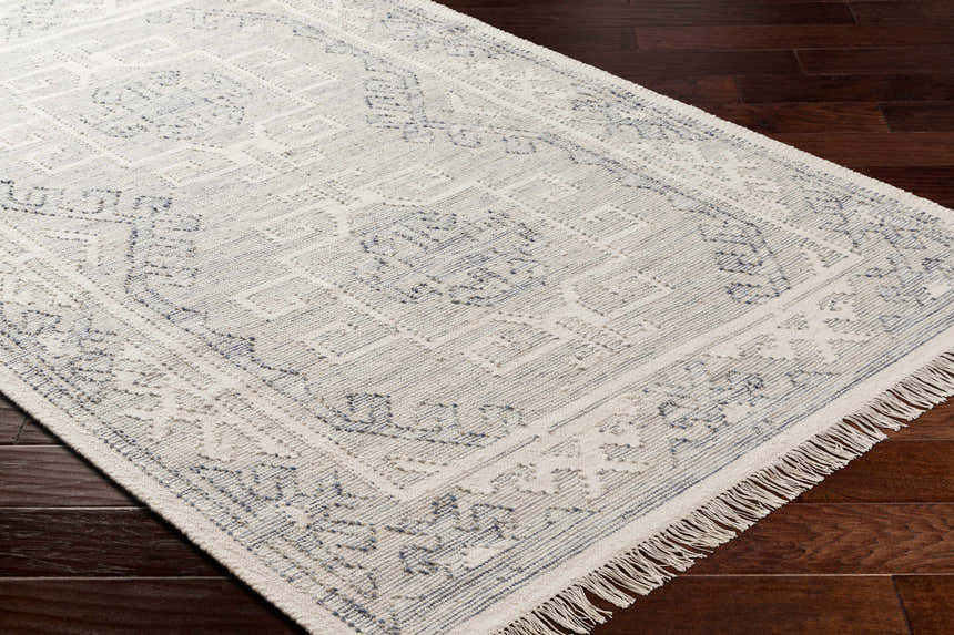 Close up of the hand woven Anund Area Rug. Beige wool rug with dark gray geometric pattern and fringe. Corner