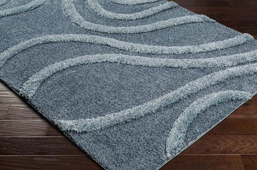 Close-up of the Taluya Area Rug. Blue shag rug with light blue wavy stripes on dark wood flooring. Contemporary style. Corner