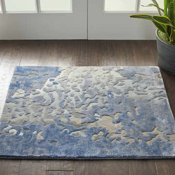 Entryway with a Nourison Prismatic Denim rug, featuring a blue abstract pattern on dark wood floors. Plant and white doors.