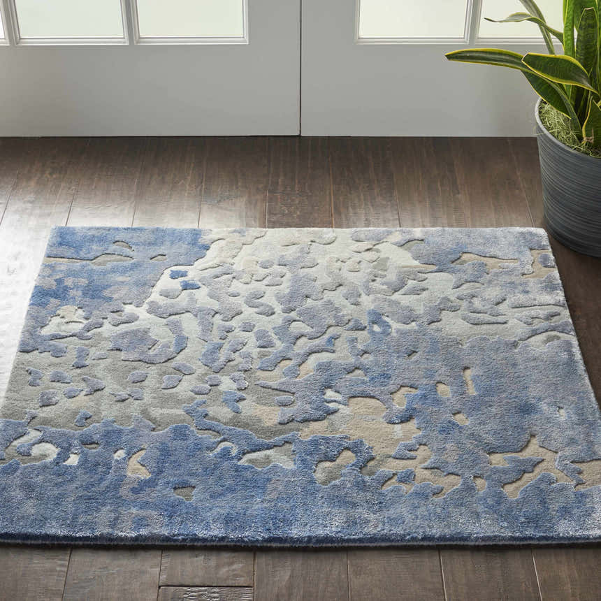 Entryway with a Nourison Prismatic Denim rug, featuring a blue abstract pattern on dark wood floors. Plant and white doors.