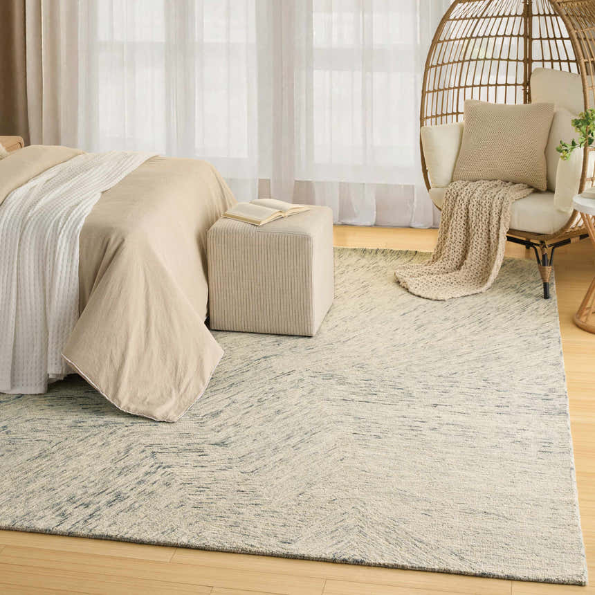Neutral bedroom scene with a DKNY Pure Angles Grey rug, bed, ottoman, and rattan chair.