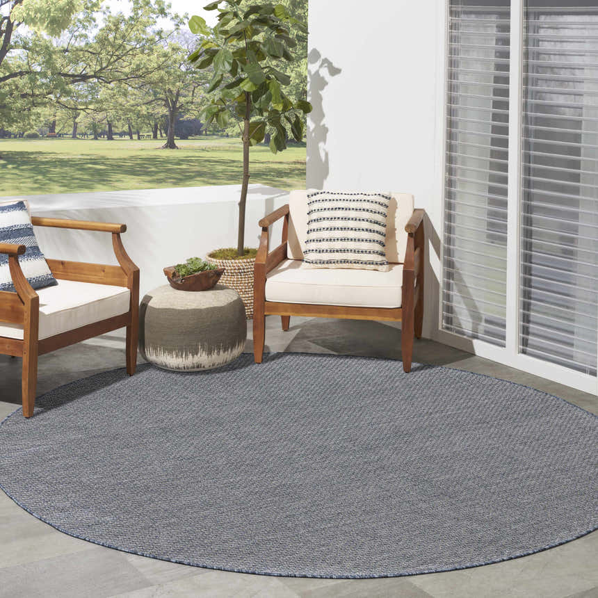 Patio scene with round Nourison Tulum Denim outdoor rug, wood chairs, and a view of a green lawn.