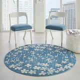 Bright room scene with a round Nourison Tranquil Turquoise rug featuring a floral pattern, white chairs, and a furry ottoman.