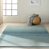 Minimalist room with aqua Calvin Klein Linear Glow rug, wood floor, round ottoman, and gray throw.