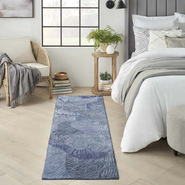 Bedroom scene with a Nourison Avra Blue Modern rug, light wood floors, rattan chair, and white bedding.