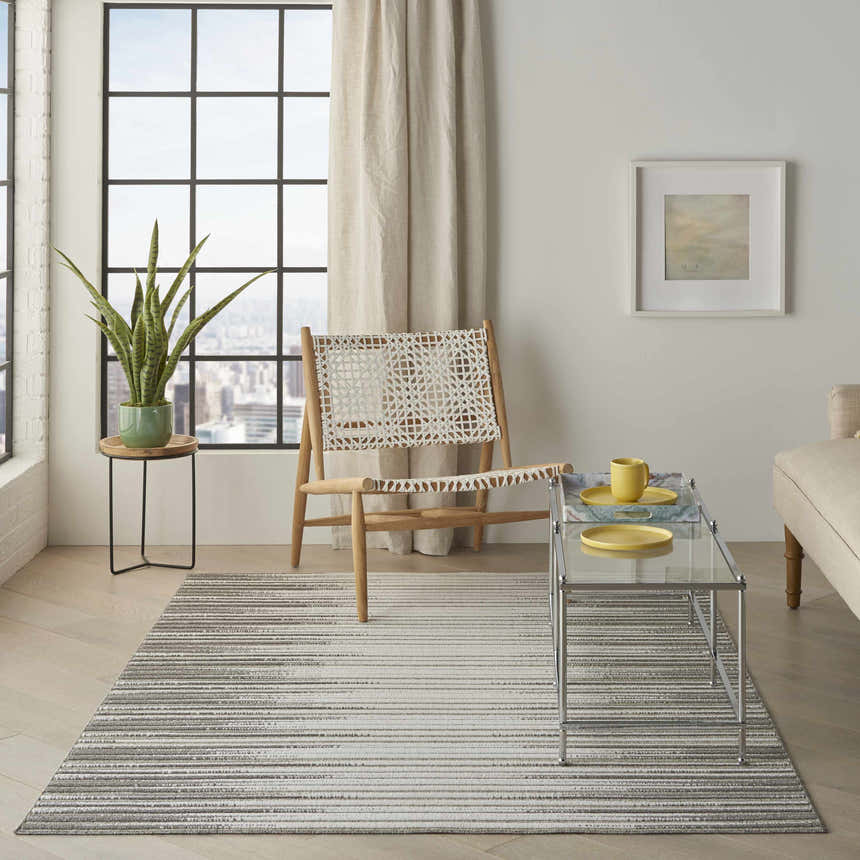 Light-filled living room with a Nourison Key Largo Light Grey rug, woven chair, glass table, and cityscape view.