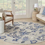 Sunroom with a round Nourison Aloha Blue Grey outdoor rug, floral pattern, wood furniture, and neutral decor.