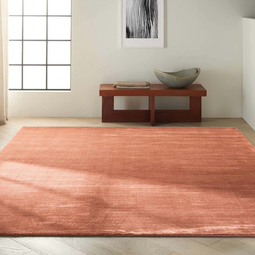 Minimalist room scene with a rust-colored Calvin Klein Home Lunar rug, wood bench, and neutral decor.