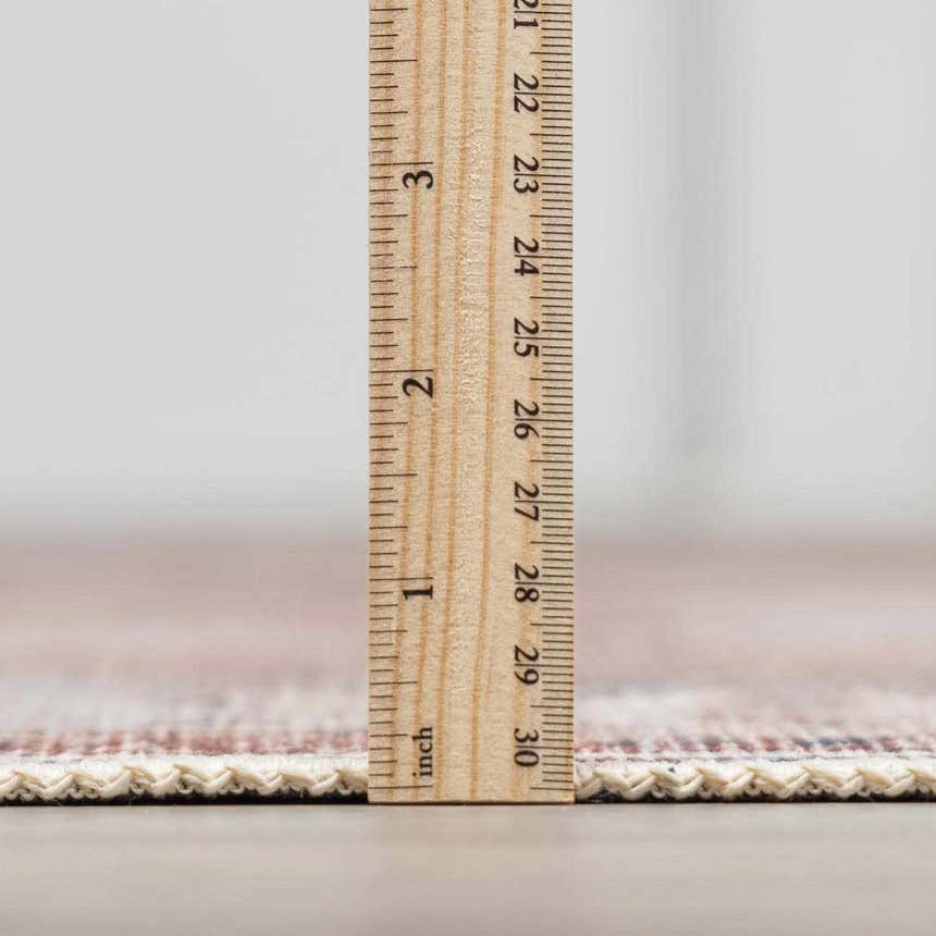 Close-up of the Arncliffe Washable Area Rug showing its pile height with a ruler. Colors: brown, ivory, and dark red. Pile