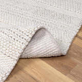 Close-up of the cream Alora Area Rug showing its chunky hand-woven texture and wool blend on a wood floor. Fold