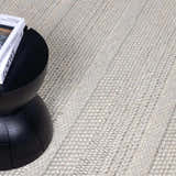Close-up of cream Alora Area Rug showing hand-woven texture next to a black side table and book. Styleshot