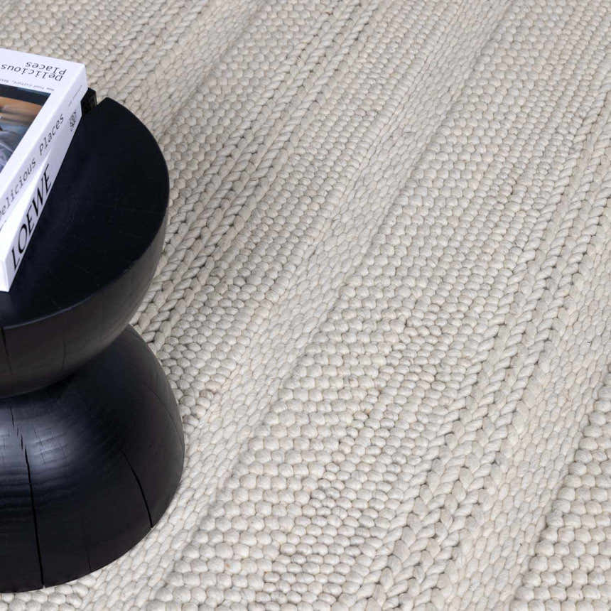 Close-up of cream Alora Area Rug showing hand-woven texture next to a black side table and book. Styleshot