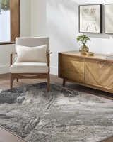 Living room scene with a beige armchair and the abstract Chaeli Area Rug in beige, black, and charcoal. Roomscene-Rectangle