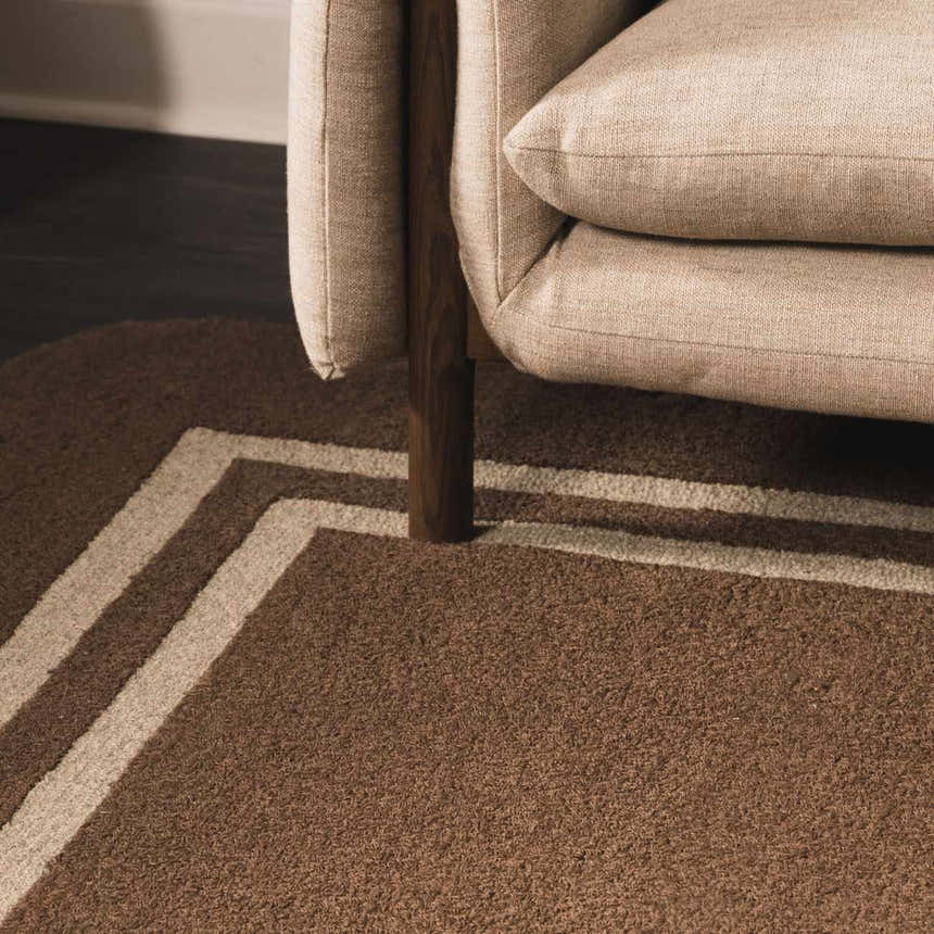 Close up of the textured Chogan Chocolate Brown Area Rug with beige border under a neutral couch. Styleshot
