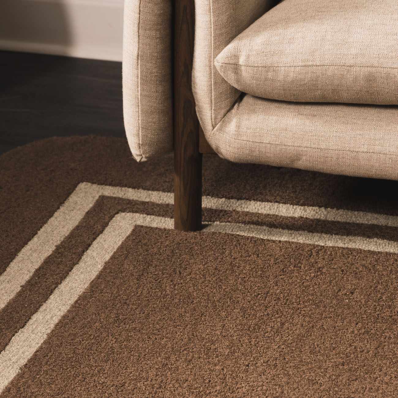 Close up of the textured Chogan Chocolate Brown Area Rug with beige border under a neutral couch. Styleshot