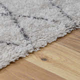 Close-up of the Flintville Moroccan Shag Area Rug. Soft, light beige shag texture with charcoal geometric pattern on wood floor. Pile
