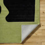 Close-up of Arvok Olive Green rug showing charcoal block design, wool texture, and folded corner on wood floor. Fold