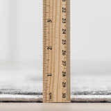 Close-up of the Littleport Luxury Fringed Faux Silk Rug showing the rug's pile height with a ruler. Gray tones. Pile