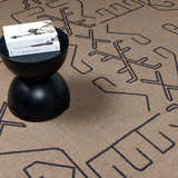 Close-up of the Hamid Washable Jutely Rug in camel with a dark brown geometric pattern and a black accent table. Styleshot