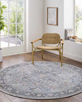 Light-filled room featuring a round Hovan Area Rug in gray tones, with a woven chair and wood floors. Roomscene-Round